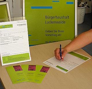 Das Foto zeigt eine Hand mit Stift beim Ausfüllen eines Formulars, im Hintergrund sieht man einen Werbeaufsteller mit Plakat zum Bürgerhaushalt. 