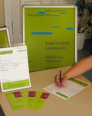 Das Foto zeigt eine Hand mit Stift beim Ausfüllen eines Formulars, im Hintergrund sieht man einen Werbeaufsteller mit Plakat zum Bürgerhaushalt. 